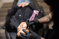 Tattooed brow artist holding tool while focusing on client’s brows. Photograph by Yucaipa branding photographer Kaitlyn Dawn Photography.