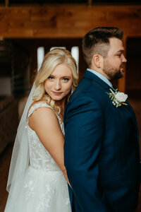Bride embraces groom from behind with soft smile during romantic indoor wedding portrait session.