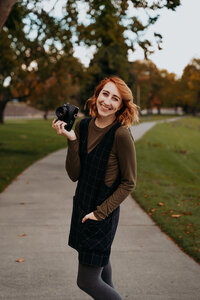 woman stands in middle of sidewalk smiling holding camera