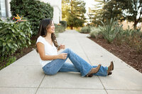 High school senior girl sitting on a sidewalk wearing jeans and boots during an outdoor portrait session by Kaitlyn Dawn Photography.