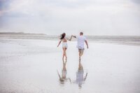 Professional photography by Nature's Child Photography: In St. Augustine, a couple runs barefoot through shallow water during a beach, wind tugging at their clothes as storm clouds gather in the distance.