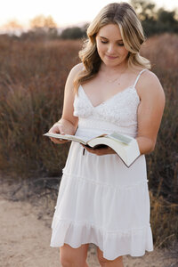 High school senior girl in a white dress reading a book in a golden hour field, outdoor senior portrait by Kaitlyn Dawn Photography.