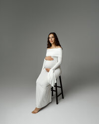Pregnant woman sitting on a stool in a long white dress, gently holding her baby bump during a Yucaipa maternity photoshoot with Kaitlyn Dawn Photography.