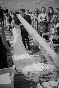 Mariée remontant l'allée au bras de son père, longue traîne de dentelle et pétales de fleurs au sol, invités assis de chaque côté, cérémonie au Belvédère du Vieux-Port de Montréal