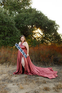 Pregnant woman wearing a flowing terracotta maternity gown standing outdoors at sunset holding ultrasound photos during a lifestyle maternity photography session captured by Kaitlyn Dawn Photography.