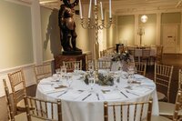 An elegant wedding reception setup featuring a round table with a white linen tablecloth, gold Chiavari chairs, and a tall gold candelabra centerpiece surrounded by greenery and glassware. The soft sage walls and classical statue in the background add a touch of old-world sophistication to the space. Captured by Phavy Photography in a true-to-color, documentary style that highlights luxury, intentional styling, and timeless wedding design. Vogue inspired