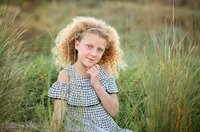 Young girl with curly blonde hair sitting in tall beach grass and smiling softly with her hand under her chin during a beach child portrait session. - A portrait experience by Nature's Child Photography.