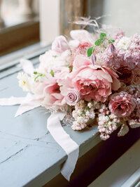 bridal bouquet featuring lilac, peonies, ranunculus, and roses in soft pink tones near a window with natural light