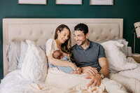 An attractive couple holding their newborn on an ivory bed against a teal wall