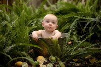 Baby sitting in a small metal tub among lush ferns and sliced citrus fruit, looking up during the splash portion of a cake smash in Mandarin. - A portrait experience by Nature's Child Photography.