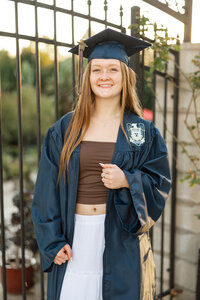 High school graduate wearing cap and gown standing by a wrought iron gate during golden hour, graduation portrait by Kaitlyn Dawn Photography.