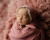 Peacefully sleeping newborn baby girl wearing a dusty rose bonnet and wrapped in a soft pink swaddle during a professional newborn photography session by Inland Empire newborn photographer Kaitlyn Dawn Photography.