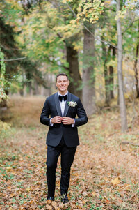 The groom stands confidently in a wooded area, smiling at the camera in his classic black tuxedo with a white floral boutonnière