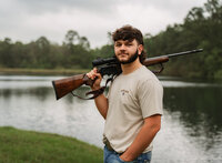 Senior portrait at Circle B Lake in Wilmer, Alabama — waist-up holding an unloaded hunting rifle
on his shoulder with the pond behind him.