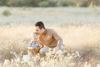 A father kneeling down next to his son in the middle of a Dallas park as he kisses his sons forehead.