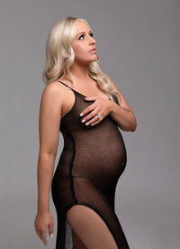 A woman in black maternity lingerie elegantly posing for a maternity photoshoot on a gray background.