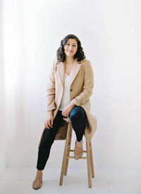A woman sitting on a wood stool looking at the camera.
