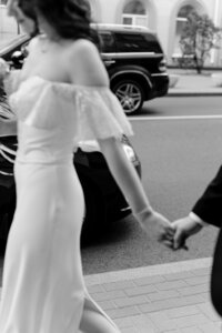 a woman wearing a white wedding dress walks down a busy street holding a man's hand