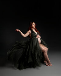 Pregnant woman with long red hair wearing a flowing dark green maternity gown while seated with the dress cascading around her and holding her baby bump during a studio maternity photography session with a dark backdrop by Kaitlyn Dawn Photography.