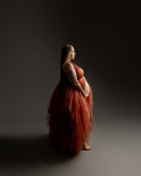 Expecting mother in a dramatic red maternity gown posing in profile during a studio maternity portrait by Kaitlyn Dawn Photography.