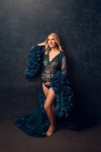 Pregnant woman wearing a dark teal lace bodysuit and ruffled robe, posing barefoot in a Waukesha photography studio with a rich textured backdrop and confident smile.