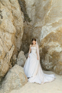 Bride looks down at her gown as she stands in front of large rock formation on Montara Beach. Photo by Jordan Katz.