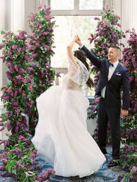 groom holding bride’s hand as she twirls during ceremony surrounded by lilac florals