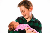 A backlit photo of a boy holding his newborn sister. He smiles down at her wearing a green plaid shirt. She is in a purple wrap, lying in his arms.