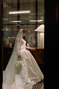 Bride in a satin gown and long veil standing by a lamp inside a luxury Huntsville hotel suite.