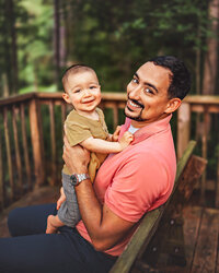 Father smiling while holding his baby on a wooded lakeside dock during outdoor family portrait session