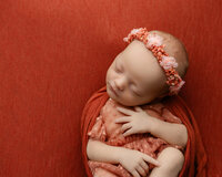 Sleeping newborn baby girl wearing a floral headband and wrapped in a deep red swaddle during a studio newborn photography portrait with newborn photographer Kaitlyn Dawn Photography.