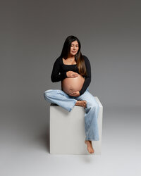 Pregnant mom sitting on a studio block in a black top and jeans, gently holding her belly during a maternity session with Kaitlyn Dawn Photography in Yucaipa.