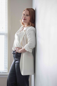 Woman in a cream blazer and black trousers leaning against a white wall beside a window, one hand on her hip.
