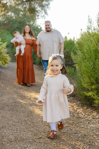Young girl walking toward the camera on a nature trail while parents follow behind holding a baby, golden hour family session captured by Kaitlyn Dawn Photography, lifestyle family photographer.