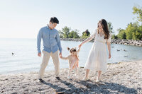 A couple holds hands with a young child, helping her swing as they stroll along a Toronto pebbly beach. With trees and a calm body of water in the background, it's the perfect scene for summer family photography.