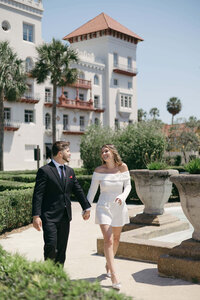 A joyful couple walks hand-in-hand outside The Lightner Museum in St. Augustine, Florida. She wears a white off-the-shoulder mini dress, and he’s in a sharp black suit with a red pocket square. Captured in a candid, documentary photography style by Phavy Photography—a St. Augustine engagement photographer specializing in luxury, architecture-inspired sessions filled with real connection and timeless charm.