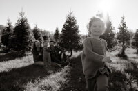 A family sits in the grass amongst the balsam trees for photos. One of the daughters runs towards the camera while the family smiles in the background.