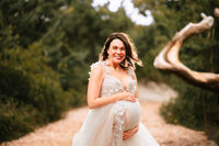 A pregnant woman in a tulle dress smiles with her hands above and below her baby bump. She stands in a wooded path and the sun is peaking down from above.