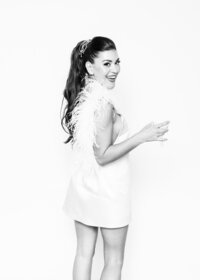 Luxury fine art portrait from Luxe Portraiture LV featuring a woman in a satin sleeveless outfit with feathered boa, high ponytail hairstyle, and decorative accessories, holding a cocktail glass and smiling in a black‑and‑white fashion‑inspired photograph that highlights elegance and glamour.