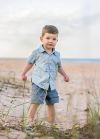 Toddler boy in a light blue button-down shirt and blue shorts walking along the sandy edge of the dunes during a beach child portrait session. (Photographed by Nature's Child Photography)