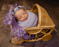 Sleeping newborn baby girl wrapped in a lavender swaddle and posed in a vintage wicker stroller prop during a studio session with newborn photographer Kaitlyn Dawn Photography.
