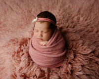 Sleeping newborn baby girl wrapped in a dusty rose swaddle and wearing a delicate headband while posed on a soft pink fur blanket during a studio newborn photography session by Kaitlyn Dawn Photography.