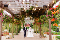 A bride and groom hold hands under a flower arbor that has bistro lights at Brookside Event Venue.