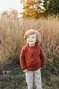 A young child with curly hair, wearing a red sweater and beige pants, smiles amidst a field of tall grass and autumn trees—capturing the essence of Toronto family photography.