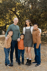 Family of five standing together outdoors surrounded by trees and golden light, coordinated earth-tone outfits captured by Kaitlyn Dawn Photography, professional outdoor family portrait photographer.