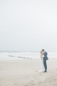 the couple holding each other on the sandy shore, with waves crashing behind them