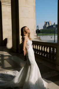 Bride in a fitted lace wedding gown standing in warm sunlight on a Paris terrace, with classical architecture and city views in the background.
