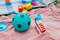 A small blue soccer ball, a toy truck, and colorful building blocks are scattered on a patterned red blanket and a sandy surface, with a blue checkered blanket in the background.