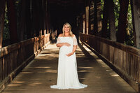 A pregnant woman in a white dress stands in the middle of a covered bridge. She holds her bump with both hands and smiles brightly.