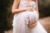 A close picture of a pregnant woman holding her baby bump. She wears a tulle dress and holds her bump with one hand above and one hand below it.